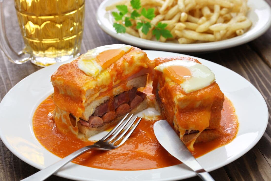 A rich Francesinha sandwich with sausage, ham, cheese, and a fresh orange sauce topped with an egg