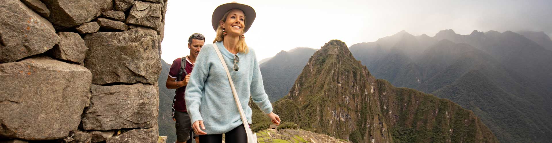 A blonde woman with a blue sweater walk in front of a man in a red t-shirt, with Machu Picchu in the background